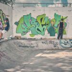 Twee mensen zijn in een met graffiti bedekt skatepark. De een rijdt op een step op de rand van een lege betonnen bak, terwijl de ander vlakbij staat en naar kleurrijke graffiti op de muur kijkt. De scène is buiten op een zonnige dag.