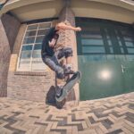 Een skateboarder in een zwart T-shirt en camouflagebroek voert een truc uit in de lucht op een stenen stoep voor een gebouw met grote groene deuren en ramen.