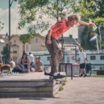 Een jongere in een rood T-shirt en bruine broek skateboardt van een houten richel in een park, terwijl mensen en een hond vlakbij zitten bij een fontein op een zonnige dag. Bomen en gebouwen zijn zichtbaar op de achtergrond.