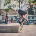Een man met een gestreept shirt en een zwarte korte broek doet een skateboardtruc op de rand van een houten platform in een park, terwijl mensen in de buurt zitten te kijken. Bomen en gebouwen zijn zichtbaar op de achtergrond.