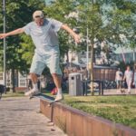 Een jongeman in een licht shirt en pet skateboardt op de rand van een lage betonnen richel in een zonnig stadspark. Op de achtergrond lopen en fietsen mensen tussen bomen en gebouwen.