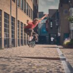 Een persoon met een rood shirt en een bruine broek skateboardt, midden op de sprong, op een met bakstenen geplaveide straat tussen hoge gebouwen op een zonnige dag. De omgeving is stedelijk, met glazen ramen en wat groen zichtbaar.