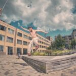 Een shirtloze skateboarder met een pet achterstevoren voert een truc uit op de rand van een verhoogd houten platform in de buurt van fonteinen op een stedelijk plein, met bakstenen gebouwen en een gedeeltelijk bewolkte lucht op de achtergrond.