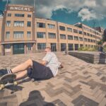 Een skateboarder met een gestreept shirt en een zwarte korte broek is van zijn skateboard gevallen op een stenen stoep voor een groot stenen gebouw met de tekst "RINGERS", terwijl een andere persoon vlakbij skateboardt.