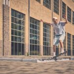 Een persoon met een witte pet, grijs T-shirt en een lichte korte broek skateboardt op een stoep voor een groot bakstenen gebouw met veel ramen en balanceert met gestrekte armen.