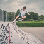 Een persoon in een grijs sweatshirt en een blauwe korte broek voert een truc uit op een skateboard bij een schans van een skatepark, die is versierd met zwarte graffiti. Op de achtergrond zijn groene struiken en bomen te zien onder een bewolkte hemel.