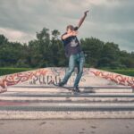 Een persoon in spijkerbroek en t-shirt voert een skateboardtruc uit op een schans bedekt met graffiti in een openlucht skatepark, met bomen en bewolkte lucht op de achtergrond.