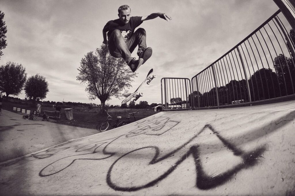 Een skateboarder voert een truc uit in de lucht boven een met graffiti bedekte schans in een openluchtskatepark, met bomen en een fiets zichtbaar op de achtergrond. De afbeelding is zwart-wit.