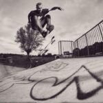 Een skateboarder voert een truc uit in de lucht boven een met graffiti bedekte schans in een openluchtskatepark, met bomen en een fiets zichtbaar op de achtergrond. De afbeelding is zwart-wit.