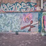 Twee mensen werken aan een kleurrijke graffiti muurschildering op een bakstenen muur; de ene hurkt links met spuitbussen, terwijl de andere rechts staat te schilderen. De muur is bedekt met levendige tags en abstracte ontwerpen.