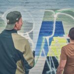 Twee mensen staan voor een kleurrijke graffitimuur; de een met een groene pet en jas, de ander met een bruin overhemd, wijzend naar het kunstwerk terwijl ze de muurschildering observeren of bespreken.