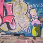 Twee mensen spuiten graffiti op een grote buitenmuur. De ene persoon staat links, terwijl de andere gehurkt kleurrijke vormen en figuren spuit. De muur is bedekt met felroze, blauwe en groene graffitikunst.