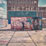 Een persoon spuit graffiti op grote, kleurrijke panelen buiten, met een gebouw met de tekst "NEDERLAND" op de achtergrond onder een gedeeltelijk bewolkte hemel.