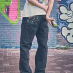 Een persoon met een grafisch t-shirt, een donkere broek en witte gympen staat op een stenen stoep met een spuitbus verf in zijn handen en kijkt naar een kleurrijke muur bedekt met graffiti. Hun gezicht is niet zichtbaar.