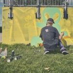 Een persoon met een zwart shirt en pet zit op gras een groene buitenaardse figuur op een gele muur te spuiten, met verfblikken in het rond. De scène is buiten bij een metalen hek onder een gedeeltelijk bewolkte hemel.