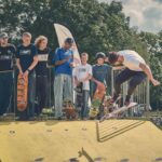 Een groep jongeren met skateboards kijkt toe hoe een skateboarder een truc uitvoert op een gele schans in een buitenpark, met bomen en fietsen op de achtergrond.