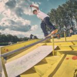 Een skateboarder slijpt midden in zijn tricks langs een gele leuning in een openluchtskatepark, met trappen eronder en een bewolkte lucht en bomen op de achtergrond. Verschillende mensen kijken toe vanuit de verte.