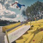 Een skateboarder in spijkerbroek en blauw shirt slijpt langs een gele leuning in een skatepark, met een paar mensen op de achtergrond onder een gedeeltelijk bewolkte hemel.