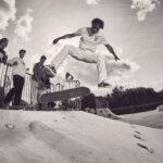 Een skateboarder voert een truc uit in de lucht in een skatepark terwijl vier mensen toekijken. De scène speelt zich buiten af onder een gedeeltelijk bewolkte hemel, vastgelegd in zwart-wit.