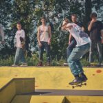 Een groep van zes mensen staat bij een skatepark te kijken naar iemand die een skateboardtruc uitvoert op een gele schans. Bomen en groen op de achtergrond en het is een zonnige dag.