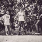 Drie mensen staan op skateboards aan de rand van een skatepark, lachend en pratend. De foto is zwart-wit en buiten genomen, met bomen op de achtergrond. Twee houden skateboards vast en één rijdt op zijn skateboard.