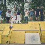 Een skateboarder doet een truc op een gele schans in een skatepark terwijl een groep mensen, sommigen met skateboards in de hand, toekijkt en op de achtergrond staat. Bomen omzomen het gebied achter hen.