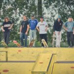 Een groep van acht skateboarders staat op een gele schans in een skatepark, houden hun skateboards vast en kijken naar beneden. Bomen en groen zijn zichtbaar op de achtergrond.