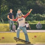 Een persoon met een rood-wit shirt en een spijkerbroek skateboardt op een gele skatehelling en balanceert op een rail. Verschillende mensen in vrijetijdskleding staan op de achtergrond toe te kijken. Bomen en groen zijn zichtbaar achter hen.