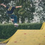 Een persoon in een zwart shirt en blauwe jeans voert een skateboardtruc uit in de lucht op een gele skatehelling in een openlucht skatepark, met bomen en struiken op de achtergrond.