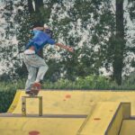 Een skateboarder met een blauw shirt en een spijkerbroek schuurt op een rail in een buiten-skatepark met gele schansen, omringd door bomen en groen.
