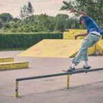 Een skateboarder in een blauw shirt en spijkerbroek balanceert op een rail in een openluchtskatepark, omgeven door hellingen en groen op een zonnige dag.