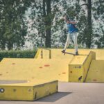 Een skateboarder in een blauw shirt en spijkerbroek doet een truc op een gele schans in een openluchtskatepark, met bomen en groen op de achtergrond.