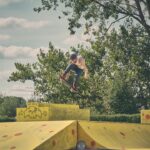 Een skateboarder in de lucht voert een truc uit over felgele schansen in een openluchtskatepark, met bomen en een bewolkte lucht op de achtergrond.