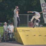 Een skateboarder voert een truc uit op een gele schans terwijl iemand in de buurt filmt. Verschillende mensen met skateboards kijken toe en op de achtergrond zijn bomen en spandoeken te zien.