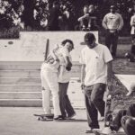 Twee mensen knuffelen in een skatepark terwijl anderen toekijken of in de buurt zitten. Skateboards zijn zichtbaar en de sfeer is ongedwongen en vriendelijk. De foto is in zwart-wit.