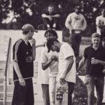 Een groep mensen staat samen buiten bij een trap, lachend en pratend. Eén persoon houdt een doos vast en een ander bedekt lachend zijn mond. Anderen op de achtergrond kijken toe of maken een praatje, waardoor een ontspannen, sociale sfeer ontstaat.