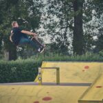 Een skateboarder in de lucht voert een truc uit over een gele schans in een buiten-skatepark, omringd door groen en bomen op de achtergrond.