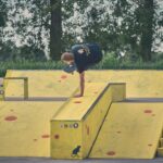 Een skateboarder balanceert op één hand op een gele schans met rode vlekken in een skatepark, omringd door groen en bomen op de achtergrond.
