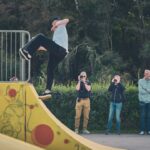 Een skateboarder in een lichtblauw shirt voert een truc uit op een gele schans met rode vlekken terwijl drie fotografen en drie toeschouwers toekijken in een parkachtige omgeving. Bomen en groen op de achtergrond.