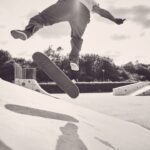 Een persoon voert een truc uit op een skateboard in een openlucht skatepark, midden in de lucht met het skateboard onder hem. De scène is zwart-wit.