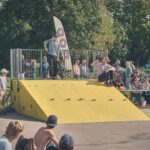 Een skateboarder doet een truc op een gele schans in een park terwijl een menigte toekijkt. Sommige mensen zitten op bankjes, anderen staan in de buurt; bomen en fietsen staan op de achtergrond. De scène heeft een ongedwongen sfeer.
