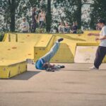 Een persoon valt met zijn hoofd voorover op de stoep in een skatepark terwijl een andere persoon nadert om te helpen. Verschillende mensen kijken toe vanaf ramps op de achtergrond. De scène speelt zich af op een zonnige dag met gele skateparkstructuren.