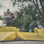 Een skateboarder in de lucht die een truc doet op een gele schans, terwijl een fotograaf de actie vastlegt. Verschillende mensen kijken toe op de achtergrond bij bomen op een bewolkte dag.