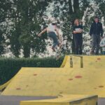 Een skateboarder voert een truc uit in de lucht in een geel skatepark terwijl drie mensen toekijken vanaf een platform. Bomen en groen zijn zichtbaar op de achtergrond.