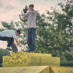 Een skateboarder voert een truc uit op een geel betonnen platform terwijl een andere persoon bovenop staat en filmt met een camera. Op de achtergrond zijn bomen en een bewolkte lucht te zien.