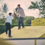 Twee mensen zijn in een skatepark; de een skateboardt een gele schans op terwijl de ander op de schans staat en naar hem uitreikt. Bomen en struiken zijn zichtbaar op de achtergrond.