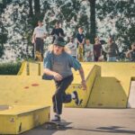Een skateboarder met lang haar, een zonnebril en een pet duwt af op zijn board in een geel skatepark, terwijl een groep mensen op de achtergrond toekijkt onder hoge bomen op een zonnige dag.