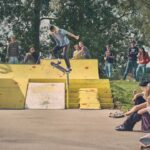 Een skateboarder voert een truc uit op een gele schans in een openlucht skatepark, gadegeslagen door een menigte mensen die rond het gebied staan en zitten. Bomen en zonlicht zijn zichtbaar op de achtergrond.