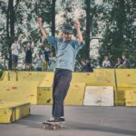 Een oudere man in een denim shirt en pet rijdt op een skateboard met opgeheven armen, lachend, voor gele schansen in een skatepark. Een groep mensen kijkt toe en ontspant op de achtergrond tussen de bomen.