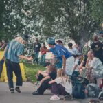 Een groep jongeren hangt rond in een skatepark, sommigen zittend en sommigen staand, met skateboards en vrijetijdskleding. Twee mensen op de voorgrond groeten elkaar hartelijk, met bomen en graffiti op de achtergrond.